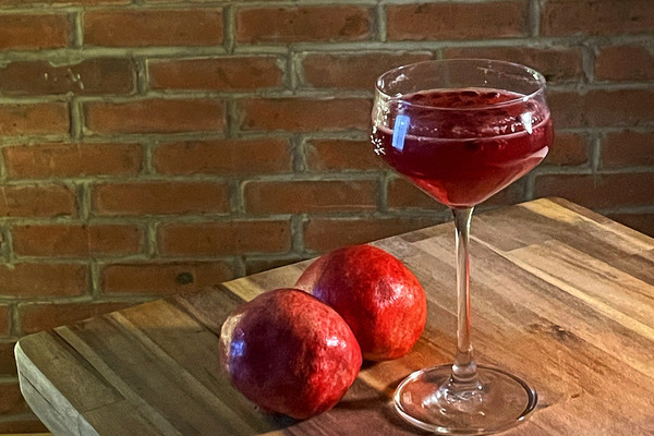 A coup fill with red liquid on a wood board, two pomegranates beside it. A brick wall in the background.