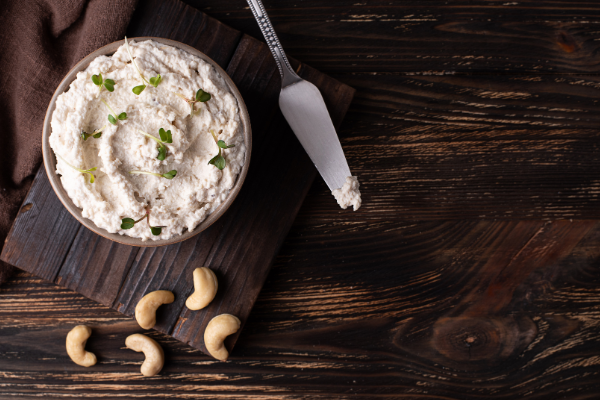 An overhead view of a glass bowl of white, spreadable cheese on a wood board, a cheese knife beside it and nuts scattered around