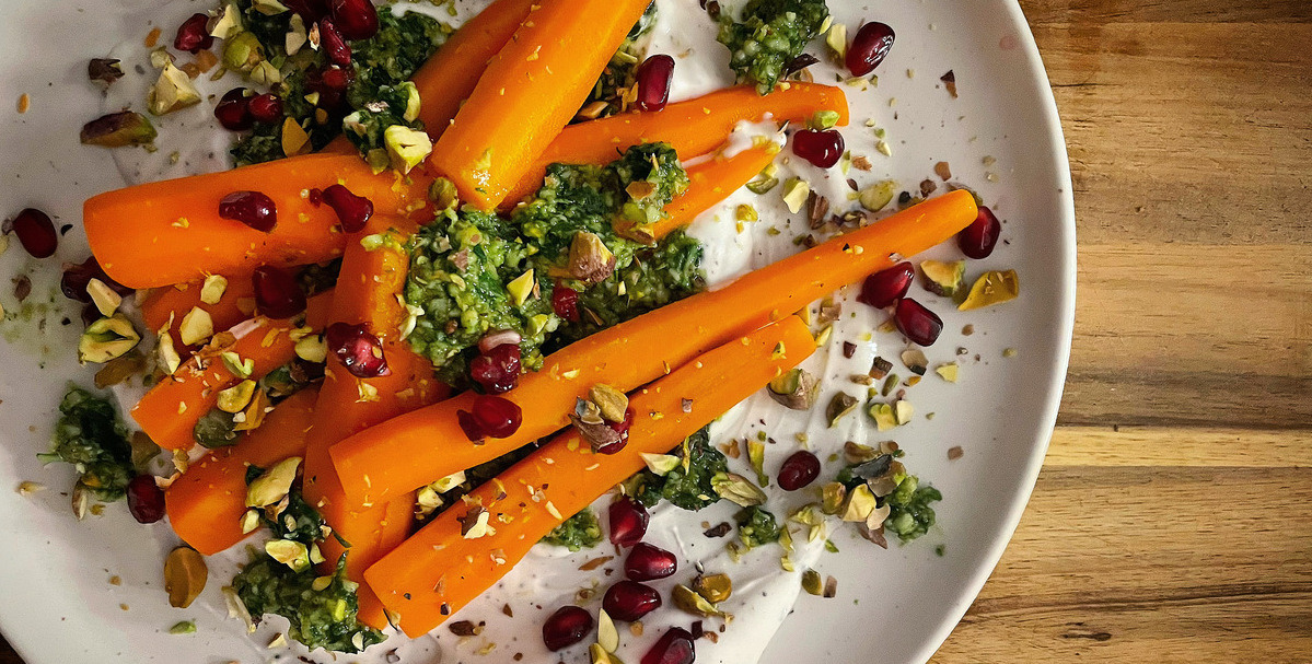 Overhead shot of a white plate on a wood board with bright orange carrots on a thick, white yogurt sauce with green pesto, red pomegranate seeds, and flecks of pistachio