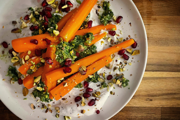Overhead shot of a white plate on a wood board with bright orange carrots on a thick, white yogurt sauce with green pesto, red pomegranate seeds, and flecks of pistachio
