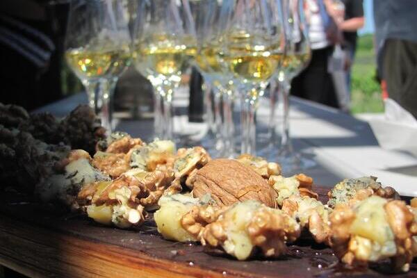 a wood board with walnuts and blue cheese, glasses of yellow icewine behind it. Blurry people in the distance