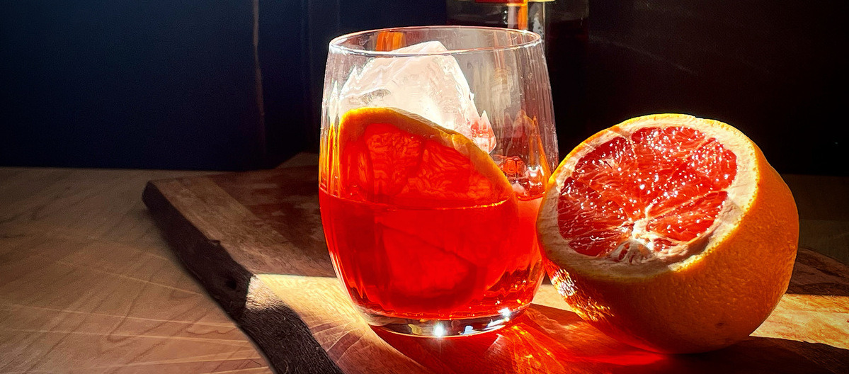 A bright red Contessa cocktail in a tumbler glass in the sunlight. A cut orange beside it.