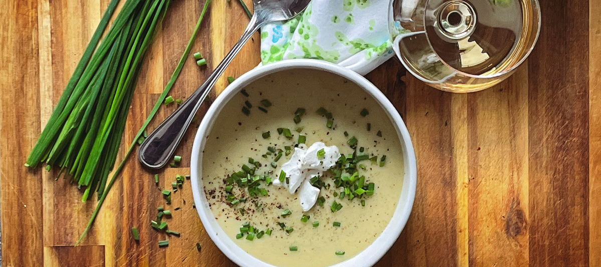 An overhead view of a white bowl filled with pale green vichyssoise on a wood board. A glass of white wine, floral napkin and chives around it.