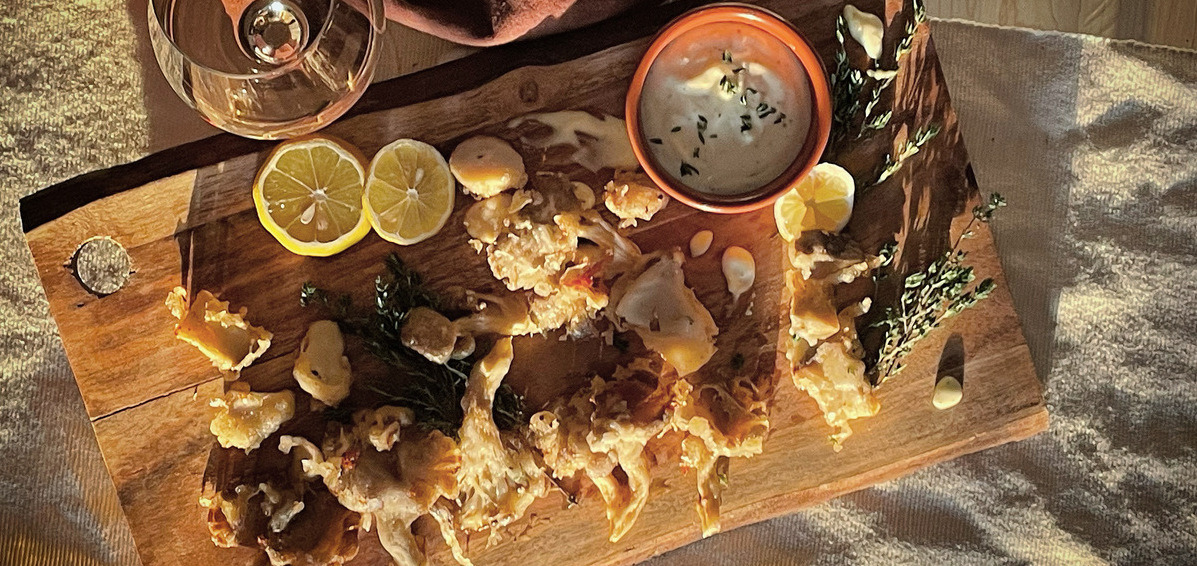 An overhead view of a pile of fried mushrooms on a wood board, a glass of white wine beside it