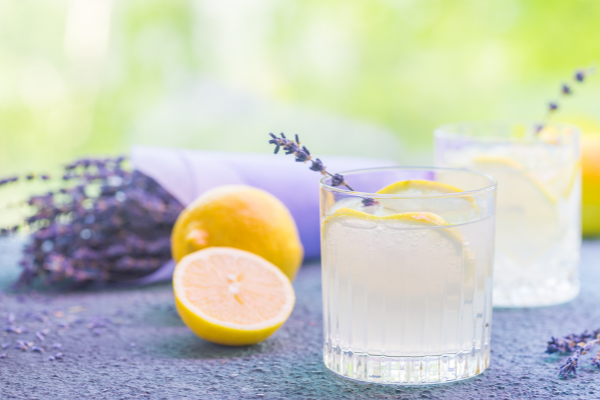 Two crystal rocks glasses with lemon slices and lavender sprigs sitting on a purple surface, a bunch of lavender in the background