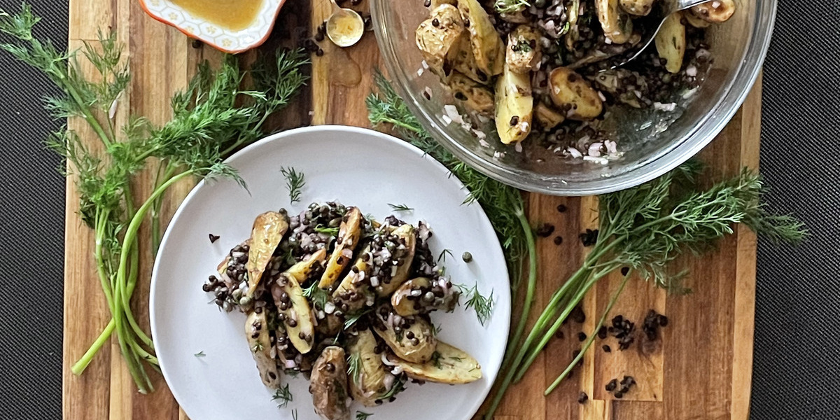 overhead view of fingerling and lentil salad on a plate beside a large serving bowl. Dill, shallot and dressing is scatter around.