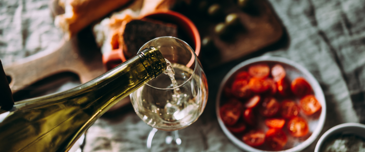 Overhead image of white wine pouring into a glass that set on a dinner table scattered with plates of food