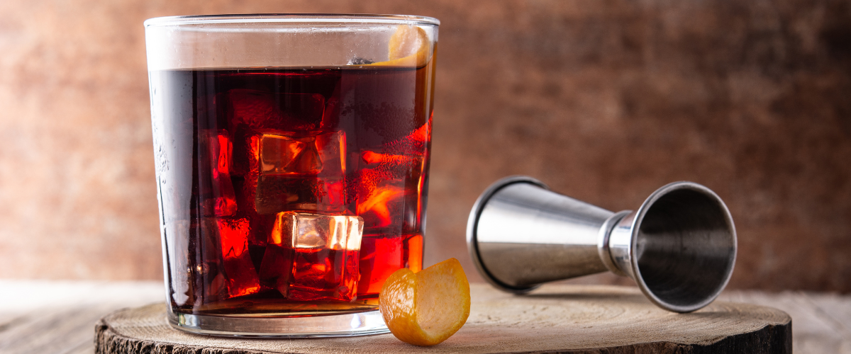 side view of a red-brown boulevardier cocktail in a rocks glass with an orange twist on a wood board
