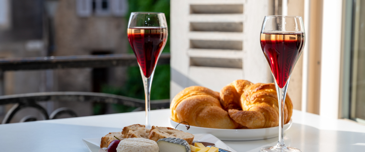 Two flutes of purple cocktails on a table in front of an open window with a European background
