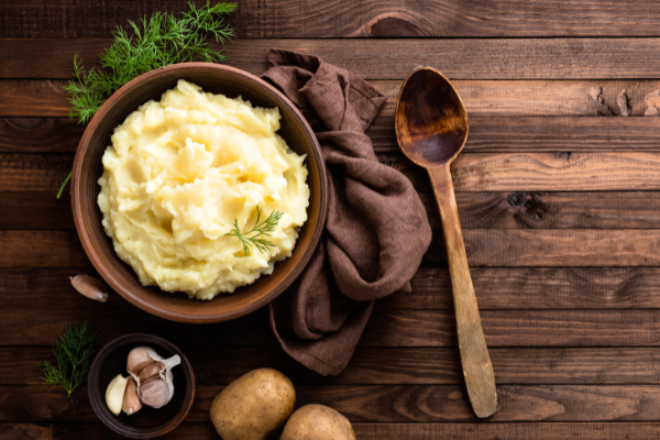 overhead view of mashed potatoes in a wood bowl on a wood counter with garlic cloves and herbs scattered around
