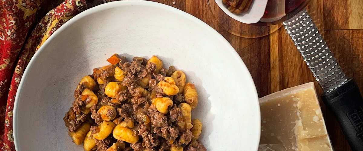 overhead view of duck porcini ragu with gnocchi in a white bowl on a wood board with a glass of red wine and parmesan cheese