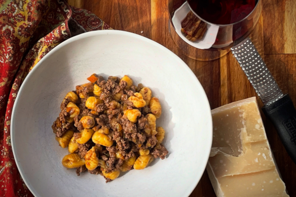 overhead view of duck porcini ragu with gnocchi in a white bowl on a wood board with a glass of red wine and parmesan cheese
