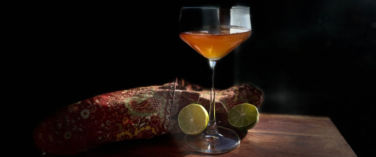 an orange cocktail in a coup against a black background; a halved lime and autumn napkin beside the glass