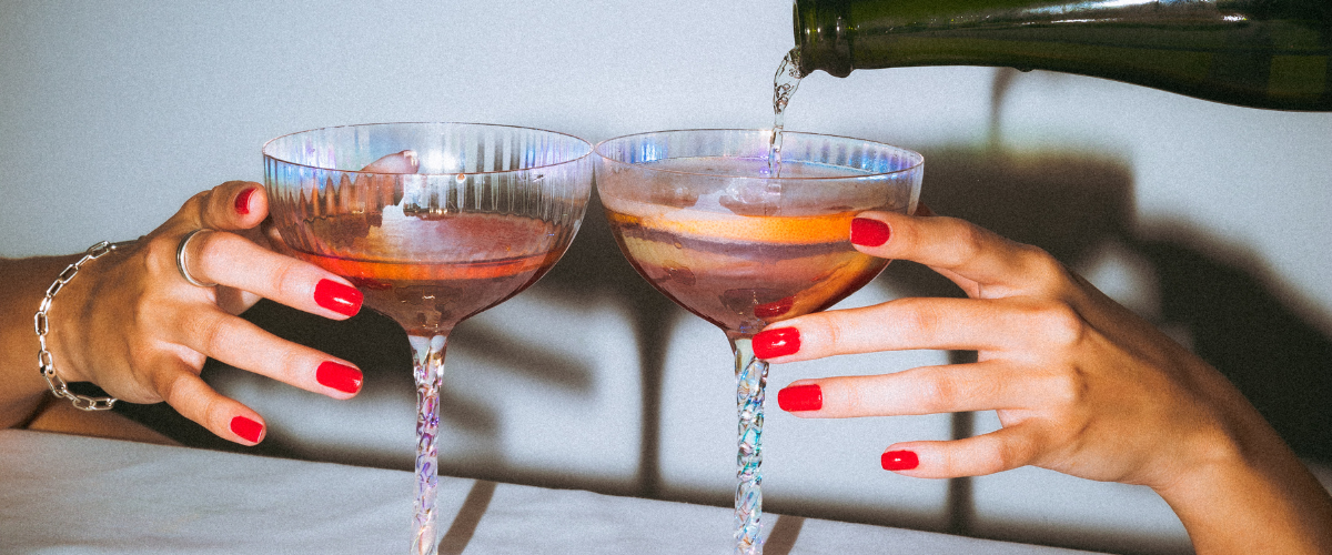 two coups with bubbly being poured into them; two hands with red nails grabbing glasses