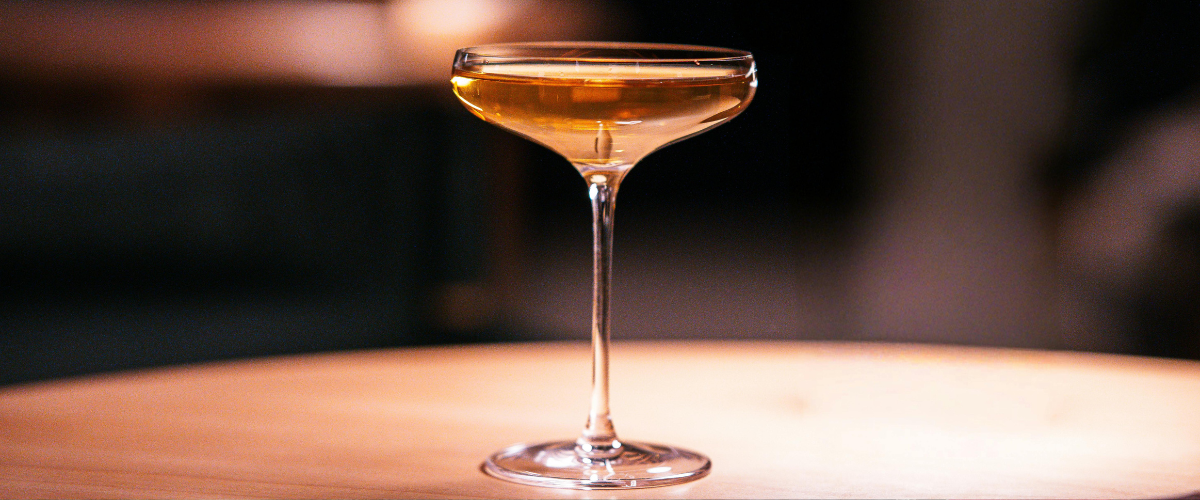 A brown cocktail in a coup on a wood table against a black background