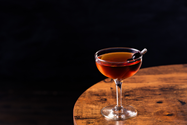 A brown cocktail in a coup on a wood table against a black background