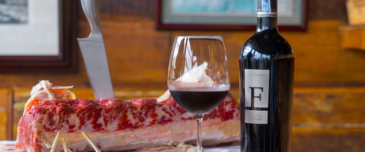 A slab of raw beef with a knife sticking out of it besdie a bottle of red wine and a glass