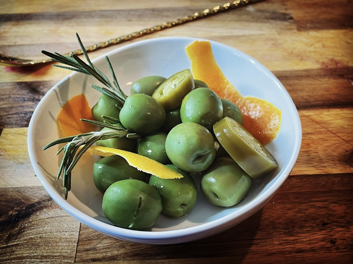 white bowl of green olives with citrus peels and herbs