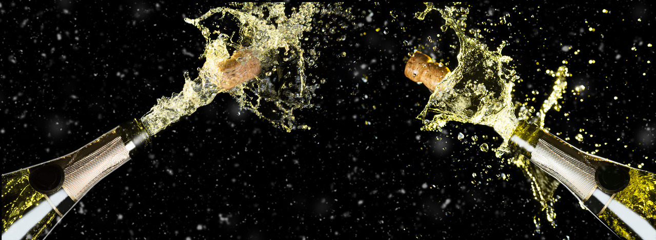 Two champagne bottles facing each other with corks blasting out and wine exploding from the bottles