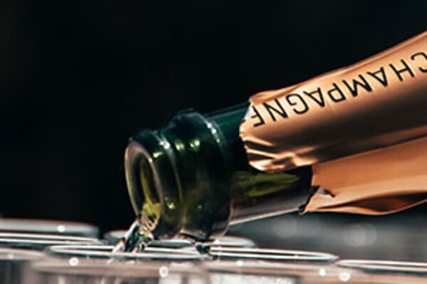 A close up of the neck of a champagne bottle pouring into the top of a champagne flute