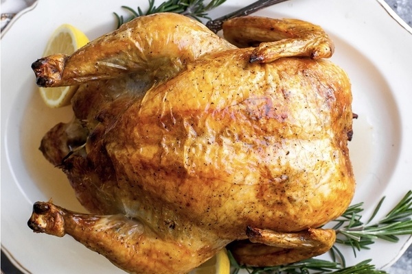 A golden roast chicken on a white platter surrounded by herbs and lemon