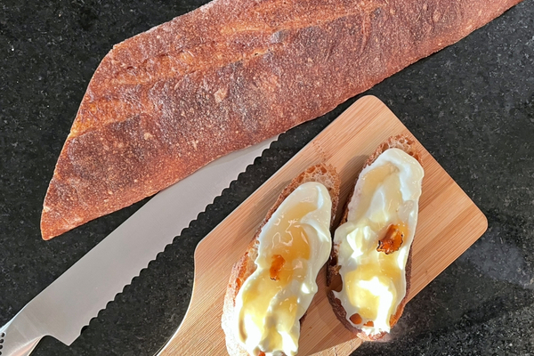 Two slices of baguette smeared with white ricotta and drizzled with truffle honey sitting on a wooden board. A bread knife and loaf are beside it.