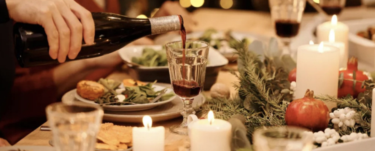 A man's hand pouring red wine into an etched crystal wine glass at a large dining table with a centre piece of candles and evergreens and plates of indistinguishable food.