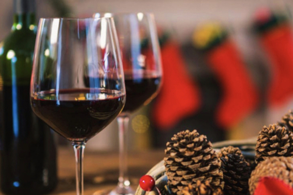 Image of two red wine glasses, a bottle of wine and a bowl of festive pinecones sitting on a wood dining table with an unlit fireplace and three red stockings in the background