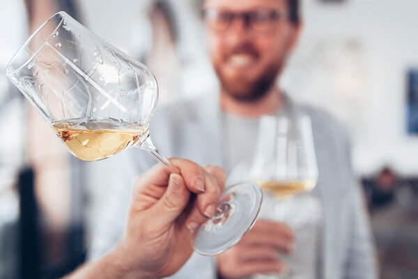 Blurry image of a man's face drinking wine and laughing in the background with a woman's hand holding a glass of white wine in the foreground