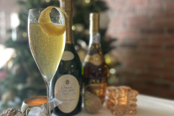 On a table with a white table cloth is a tall flute of French 75 with a lemon twist; in the background is an out-of-focus sparkling wine bottle and brandy bottle. A brick wall and christmas tree are behind the table.