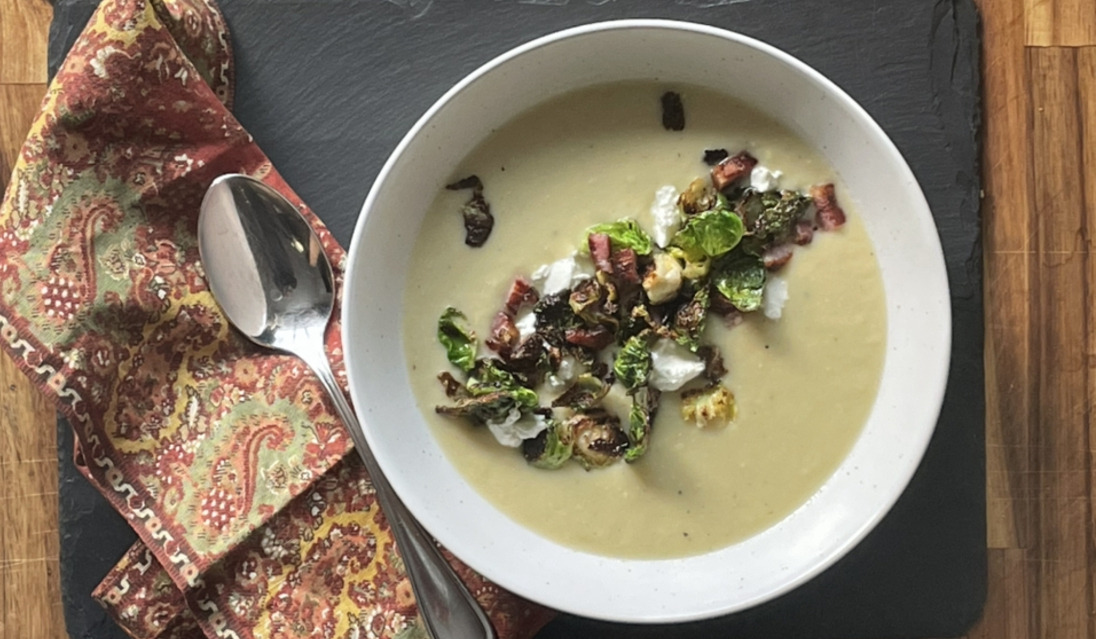 An overhead view of creamy potto soup in a white bowl, garnished with crisp brussles sprout leaves and pancetta. It sits on a black board and beside the bowl is a shiny silver spoon on a red paisley linen napkin.