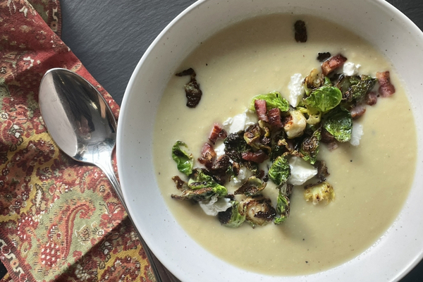 An overhead view of creamy potto soup in a white bowl, garnished with crisp brussles sprout leaves and pancetta. It sits on a black board and beside the bowl is a shiny silver spoon on a red paisley linen napkin.