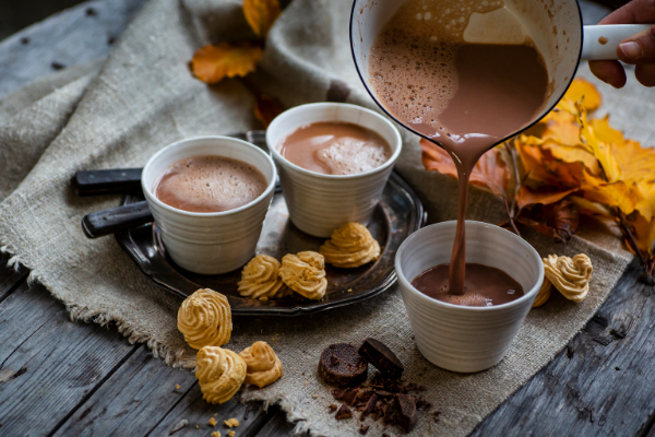 side view of a white enamel pot pouring hot chocolate into a white handleless mug, two small white cups filled with hot chocolate on a rustic board behind it.