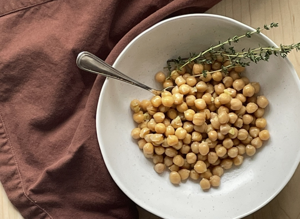 A white bowl of chickpeas with thyme scatter overtop. A brown napkin is beside the bowl.