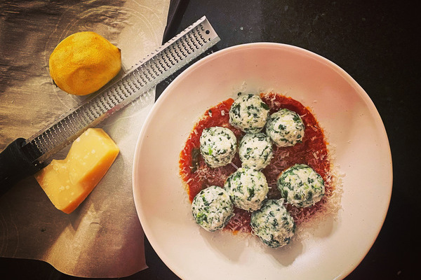 An overview picture of a white bowl with tomato sauce and eight, white, round gnudi dumplings flecked with green spinach. to the side is a baking sheet with a lemon and hunk of parmesan cheese with a microplane.