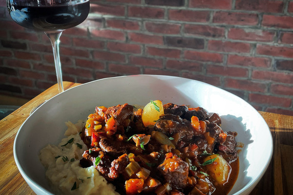 A white bowl filled with steaming boeuf bourguignon on mashed potatoes, a glass of red wine and bottle out of focus in the background