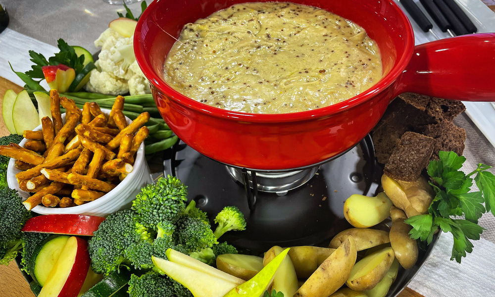 melted cheese fondue in a red bowl surrounded by various cut up vegetables, fruit and bread