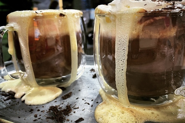 Two big glass mugs of hot chocolate, topped with whipped cream that is overflowing down the sides