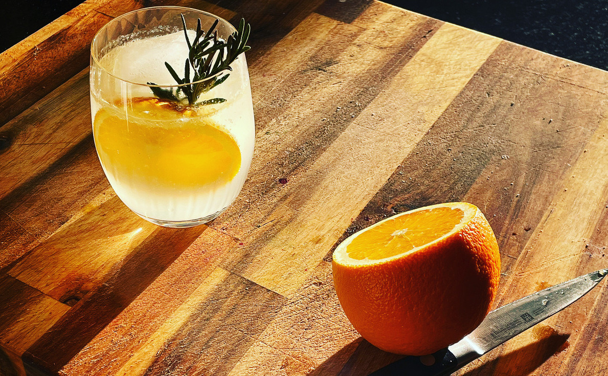A glass tumbler with clear liquid, an orange wheel and sprig of rosemary sit on a wooden cutting board, illuminated by sunlight. Beside the glass is a half cut orange and pairing knife with water drops on it.