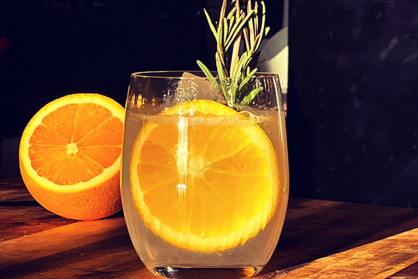 A black background with a glass tumbler filled with clear liquid and a wheel of orange and sprig of rosemary illuminated by the setting sun. In the background is a cut, half orange. Everything sits on a wooden cutting board.