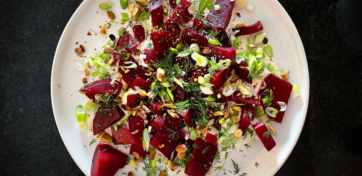 overhead view of purple beets with sliced green onion, dill and pistachio on a white circular plate on a black counter