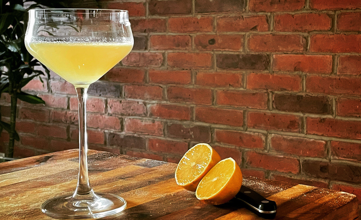 White lady cocktail of yellow liquid in a coup sitting on a wooden board with a cut lemon beside it. A brick wall is behind the drink.