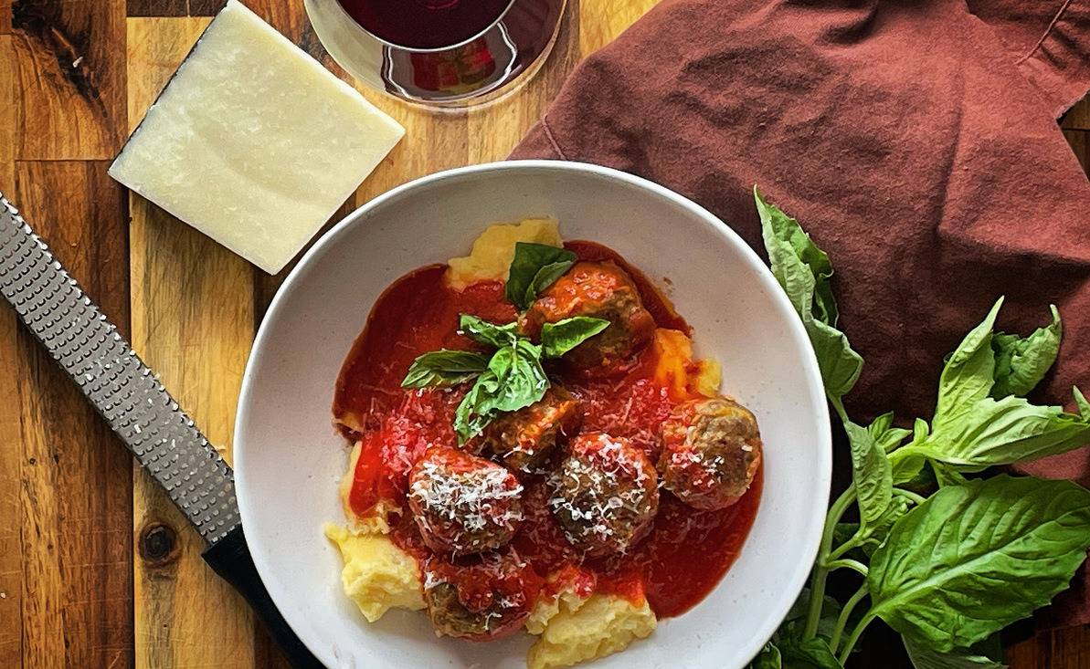 Overhead view of meatballs in red sauce in a white bowl beside a brown cloth napkin, a cluster of fresh basil and a wedge of cheese