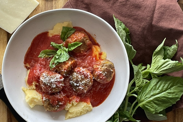 Overhead view of meatballs in red sauce in a white bowl beside a brown cloth napkin, a cluster of fresh basil and a wedge of cheese