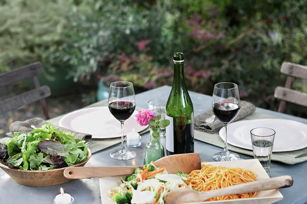 An outdoor bistro table with a plate of pasta and a bowl of salad along with a bottle of red wine and two glasses.