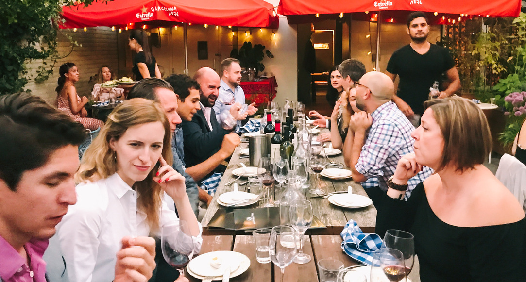 A long picnic table outside, people sit along either side, a selection of food and wine scattered across.