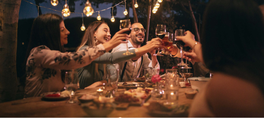 a group of people sitting around an outdoor picnic table at dusk, clinking wine glasses and laughing under string lights
