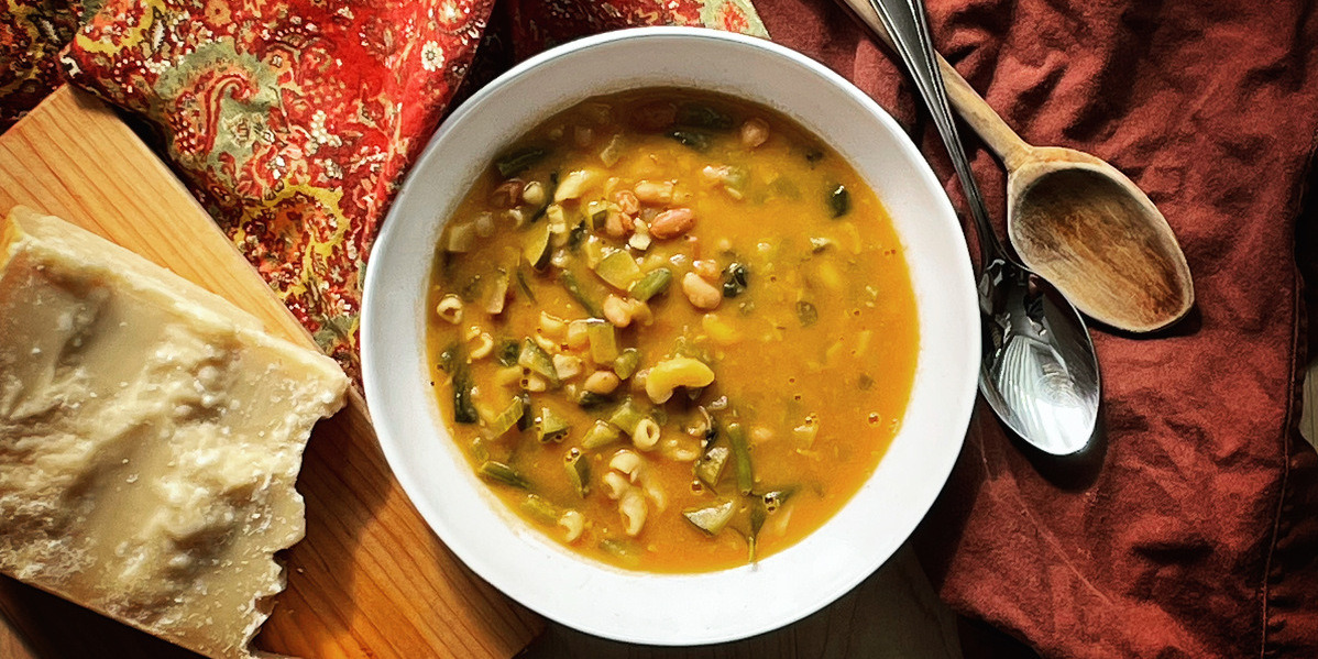 a white bowl filled with hearty minestrone. beside the bowl is a hunk of crumbly parmesan on a wood board and a rust coloured napkin with a wood spoon