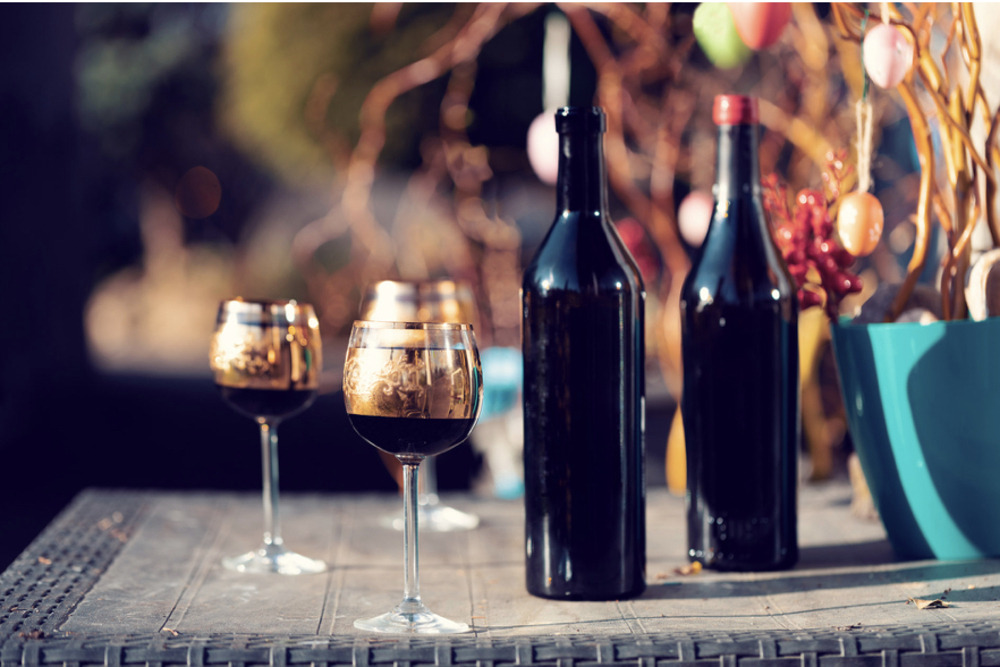 two unidentified wine bottles on a sunny, outdoor patio, with three wine glasses filled with red wine beside the. The centrepiece is a collection of branches with decorated eggs hanging from them.