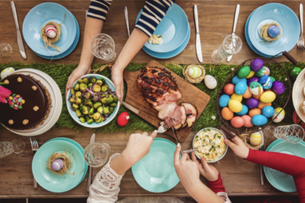 A wood dining table shown from above, set for six with turquoise blue plates, a spiral ham, basket of colourful eggs, and a chocolate cake.